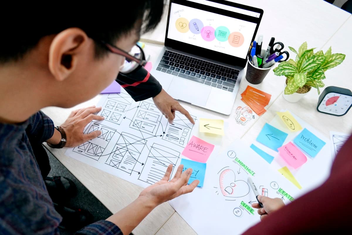 A designer reviewing hand-drawn wireframes next to a laptop showing a UX process diagram