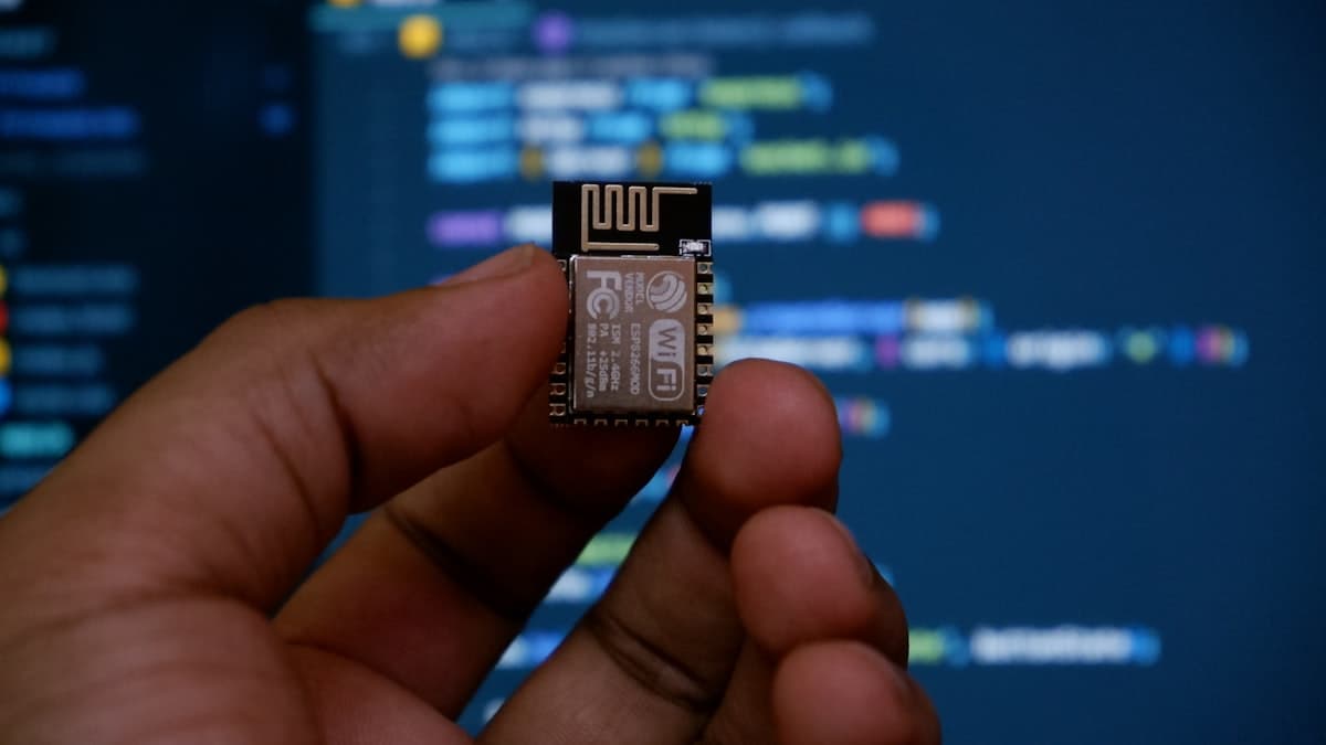 A hand holding a Wi-Fi microcontroller chip in front of a code editor